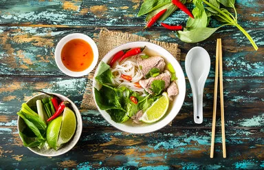 Eine vietnamesische Suppe steht auf einem hölzernen Untergrund, links daneben stehen eine Schüssel mit frischen Zuaten und eine Soße.