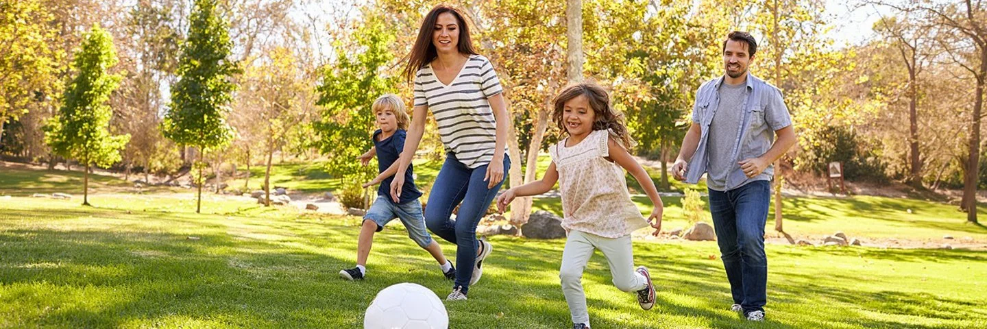Eine Familie mit zwei Kindern läuft lachend auf einem Rasen einem weißen Ball hinterher