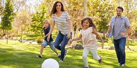 Eine Familie mit zwei Kindern läuft lachend auf einem Rasen einem weißen Ball hinterher