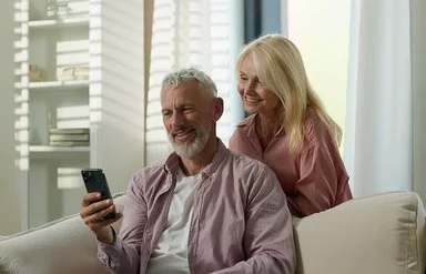 Ein Mann im fliederfarbenen Hemd und weißen T-Shirt sitzt auf einem Sofa und hält ein Handy in der Hand. Eine Frau in rosa Bluse steht lächelnd hinter ihm.