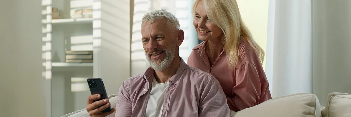 Ein Mann im fliederfarbenen Hemd und weißen T-Shirt sitzt auf einem Sofa und hält ein Handy in der Hand. Eine Frau in rosa Bluse steht lächelnd hinter ihm.