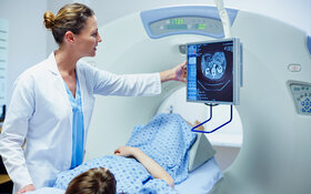 Eine Frau in einem weißen Kittel steht an einem Computer, während eine andere Frau darunter auf einer Bank liegt, die in ein CT-Gerät einfährt.