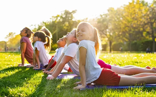 Eine Gruppe von sechs Kindern macht mit einer Trainerin zusammen Kinderyoga-Übungen im Park.