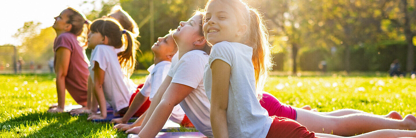 Eine Gruppe von sechs Kindern macht mit einer Trainerin zusammen Kinderyoga-Übungen im Park.