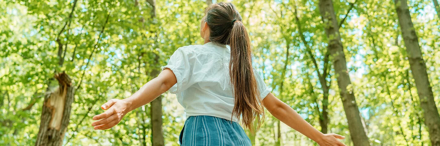 Eine junge Frau steht mit geöffneten Armen im Wald und blickt hinauf in den Himmel.