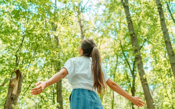 Eine junge Frau steht mit geöffneten Armen im Wald und blickt hinauf in den Himmel.