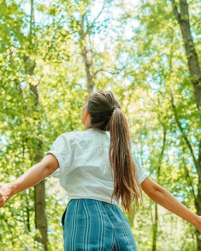 Eine junge Frau steht mit geöffneten Armen im Wald und blickt hinauf in den Himmel.