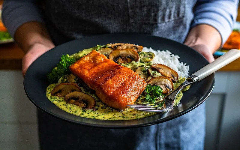 Eine Person in einer Schürze, deren Gesicht nicht im Bild ist, hält einen Teller, auf dem sich ein gebratenes Lachsfilet mit Reis, Pilzen, Spinat und Sauce befindet.