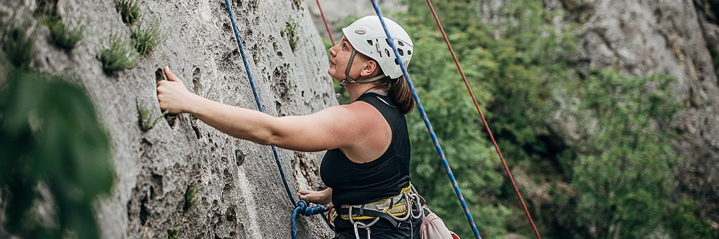 Eine Frau mit Steighelm, die mit Seilen und Gurten gesichert ist, klettert eine natürliche Felswand hinauf.