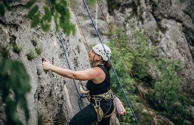 Eine Frau mit Steighelm, die mit Seilen und Gurten gesichert ist, klettert eine natürliche Felswand hinauf.