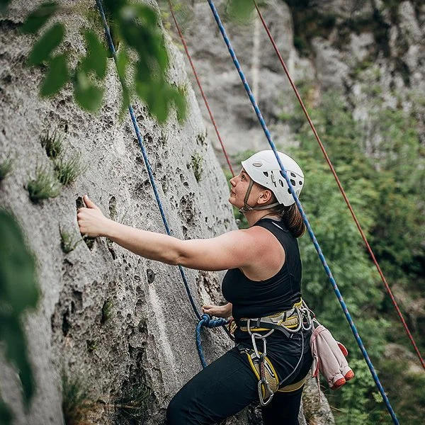 Eine Frau mit Steighelm, die mit Seilen und Gurten gesichert ist, klettert eine natürliche Felswand hinauf.