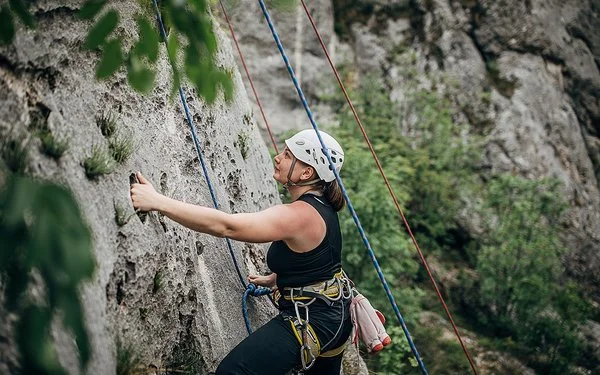 Eine Frau mit Steighelm, die mit Seilen und Gurten gesichert ist, klettert eine natürliche Felswand hinauf.
