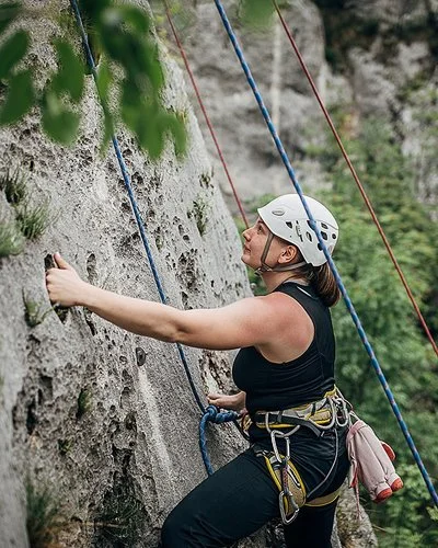 Eine Frau mit Steighelm, die mit Seilen und Gurten gesichert ist, klettert eine natürliche Felswand hinauf.