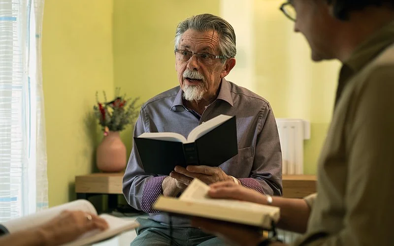 In einem Raum sitzen drei Personen. Von der Person links sind nur die Hände erkennbar, die Person rechts ist im Profil gezeigt. In der Mitte sitzt ein älterer Mann. Alle halten ein schwarz eingebundenes Buch in den Händen. Der Mann schaut die linke Person an und spricht mit ihr.
