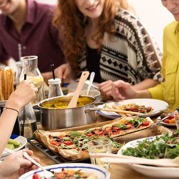 Eine Gruppe junger Menschen sitzt an einem reich gedeckten Tisch auf dem Gemüsesticks, eine Schüssel Salat, ein Topf mit Reis und eine vegetarische Pizza stehen.