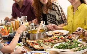 Eine Gruppe junger Menschen sitzt an einem reich gedeckten Tisch auf dem Gemüsesticks, eine Schüssel Salat, ein Topf mit Reis und eine vegetarische Pizza stehen.