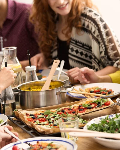 Eine Gruppe junger Menschen sitzt an einem reich gedeckten Tisch auf dem Gemüsesticks, eine Schüssel Salat, ein Topf mit Reis und eine vegetarische Pizza stehen.