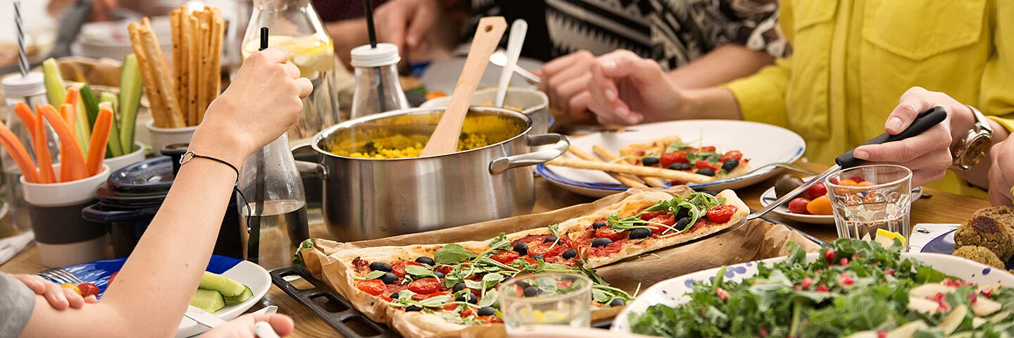 Eine Gruppe junger Menschen sitzt an einem reich gedeckten Tisch auf dem Gemüsesticks, eine Schüssel Salat, ein Topf mit Reis und eine vegetarische Pizza stehen.