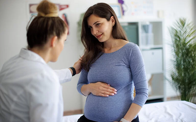Eine schwangere Frau sitzt bei ihrer Frauenärztin auf der Untersuchungsliege. Die Frauenärztin spricht mit der Patientin und legt eine Hand auf deren Schulter.