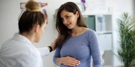 Eine schwangere Frau sitzt bei ihrer Frauenärztin auf der Untersuchungsliege. Die Frauenärztin spricht mit der Patientin und legt eine Hand auf deren Schulter.