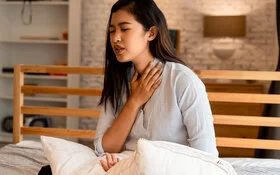Eine junge Frau sitzt aufrecht im Bett und fasst sich wegen Atemnot an den Hals.