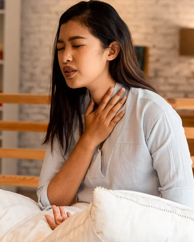 Eine junge Frau sitzt aufrecht im Bett und fasst sich wegen Atemnot an den Hals.