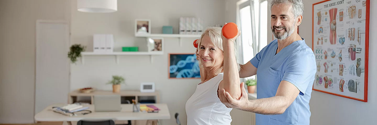 Eine ältere Frau lässt sich von einem Physiotherapeuten Übungen mit Hanteln zeigen.