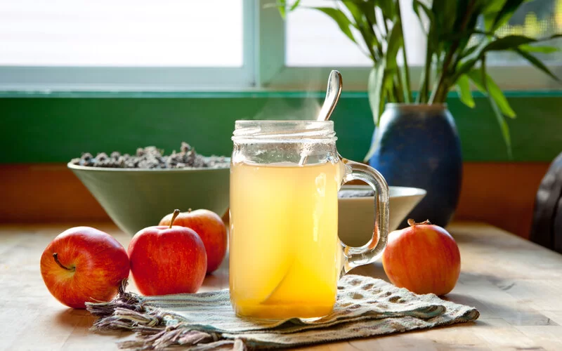 Ein Glas mit heißem Apfelessig steht auf einem Tisch, daneben Äpfel.