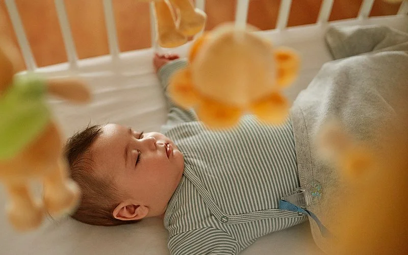 Ein Baby schläft auf dem Rücken, die Arme zur Seite gestreckt, in einem hellen Gitterbett.