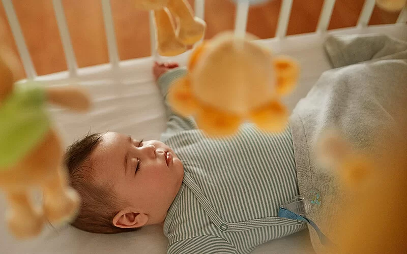 Ein Baby schläft auf dem Rücken, die Arme zur Seite gestreckt, in einem hellen Gitterbett.