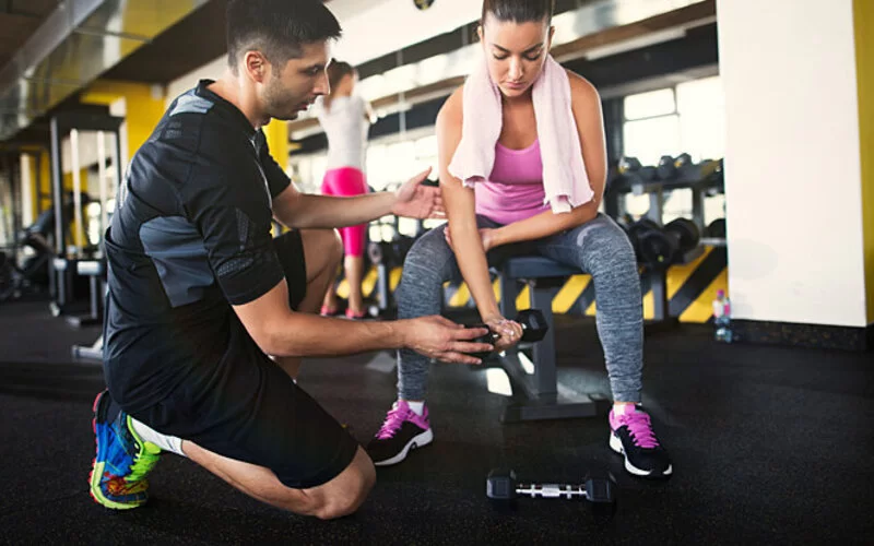 Ein Personaltrainer hilft einer jungen sportlichen Frau beim Training mit Hanteln im Fitnessstudio.