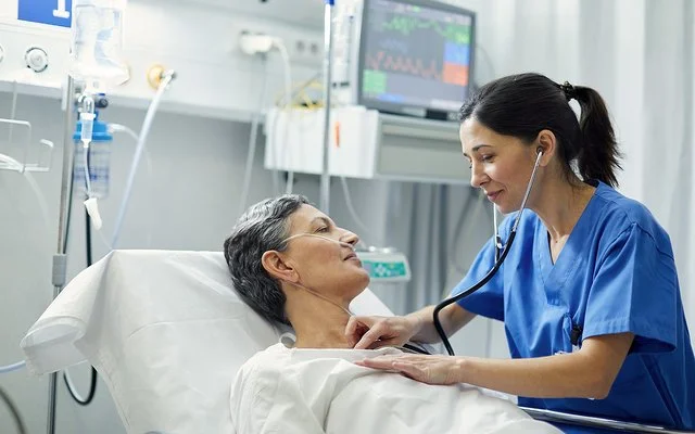 Eine Krankenschwester horcht mit dem Stethoskop die Brust einer Patientin ab, die im Bett liegt. Die Patientin hat eine Nasensonde, im Hintergrund sind Infusionen und ein Monitor zu sehen.