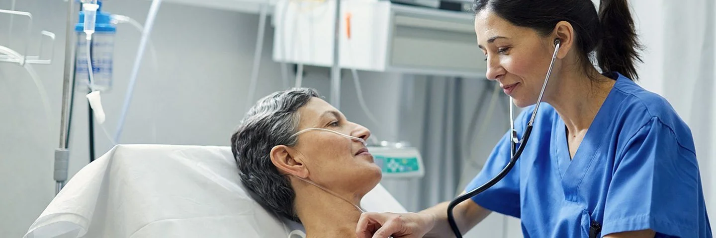 Eine Krankenschwester horcht mit dem Stethoskop die Brust einer Patientin ab, die im Bett liegt. Die Patientin hat eine Nasensonde, im Hintergrund sind Infusionen und ein Monitor zu sehen.