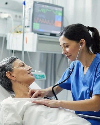 Eine Krankenschwester horcht mit dem Stethoskop die Brust einer Patientin ab, die im Bett liegt. Die Patientin hat eine Nasensonde, im Hintergrund sind Infusionen und ein Monitor zu sehen.