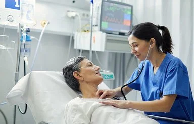 Eine Krankenschwester horcht mit dem Stethoskop die Brust einer Patientin ab, die im Bett liegt. Die Patientin hat eine Nasensonde, im Hintergrund sind Infusionen und ein Monitor zu sehen.