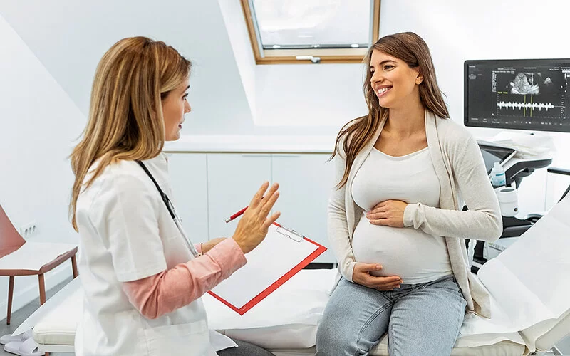 Eine schwangere Frau sitzt auf der Untersuchungsliege im Behandlungszimmer einer Gynäkologin. Sie hält sich den Bauch und folgt lächelnd den Ausführungen ihrer Frauenärztin, die ihr gegenübersitzt.