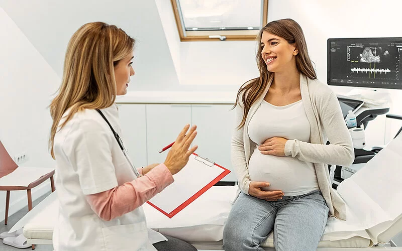 Eine schwangere Frau sitzt auf der Untersuchungsliege im Behandlungszimmer einer Gynäkologin. Sie hält sich den Bauch und folgt lächelnd den Ausführungen ihrer Frauenärztin, die ihr gegenübersitzt.