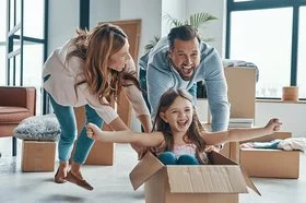 Junge Eltern schieben ihre Tochter in einem Umzugsskarton zum Spaß über den Boden.