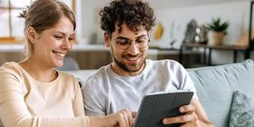 Ein junges Paar sitzt auf einem Sofa in einer hell eingerichteten Dachwohnung. Der Mann hält ein Tablet in seinen Händen, die Frau tippt aufs Display. Beide lächeln fröhlich. Im Hintergrund sind mehrere Grünpflanzen zu erkennen.