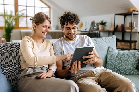 Ein junges Paar sitzt auf einem Sofa in einer hell eingerichteten Dachwohnung. Der Mann hält ein Tablet in seinen Händen, die Frau tippt aufs Display. Beide lächeln fröhlich. Im Hintergrund sind mehrere Grünpflanzen zu erkennen.
