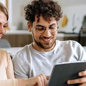Ein junges Paar sitzt auf einem Sofa in einer hell eingerichteten Dachwohnung. Der Mann hält ein Tablet in seinen Händen, die Frau tippt aufs Display. Beide lächeln fröhlich. Im Hintergrund sind mehrere Grünpflanzen zu erkennen.