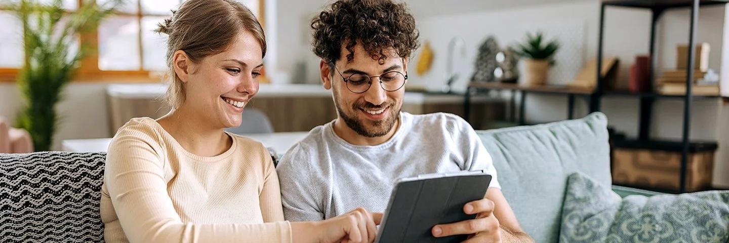 Ein junges Paar sitzt auf einem Sofa in einer hell eingerichteten Dachwohnung. Der Mann hält ein Tablet in seinen Händen, die Frau tippt aufs Display. Beide lächeln fröhlich. Im Hintergrund sind mehrere Grünpflanzen zu erkennen.