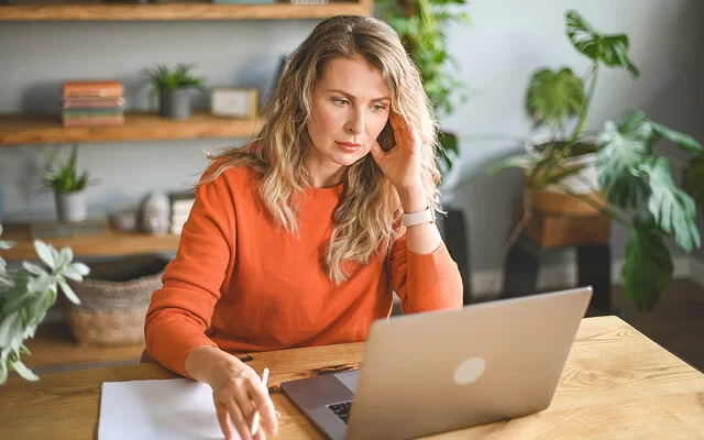 Eine Frau sitzt mit müdem Blick vor einem Laptop, den Kopf mit einer Hand gestützt.