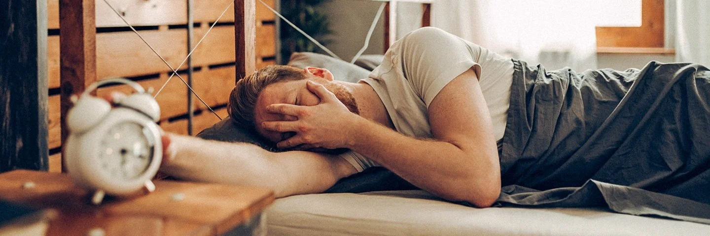 Ein Mann liegt im Bett und wurde von seinem Wecker aus dem Schlaf gerissen.