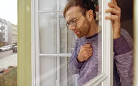 Mann mit Feinstaubbelastung steht hustend am geöffneten Fenster.