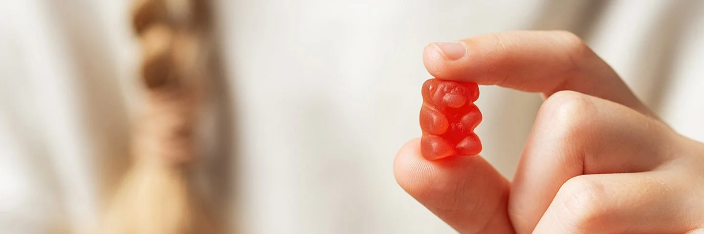 Im Vordergrund eine Kinderhand, die ein rotes Gummibärchen zwischen Zeigefinger und Daumen hält. Im Hintergrund in der Unschärfe der weiße Pullover und der Zopf eines Mädchens.