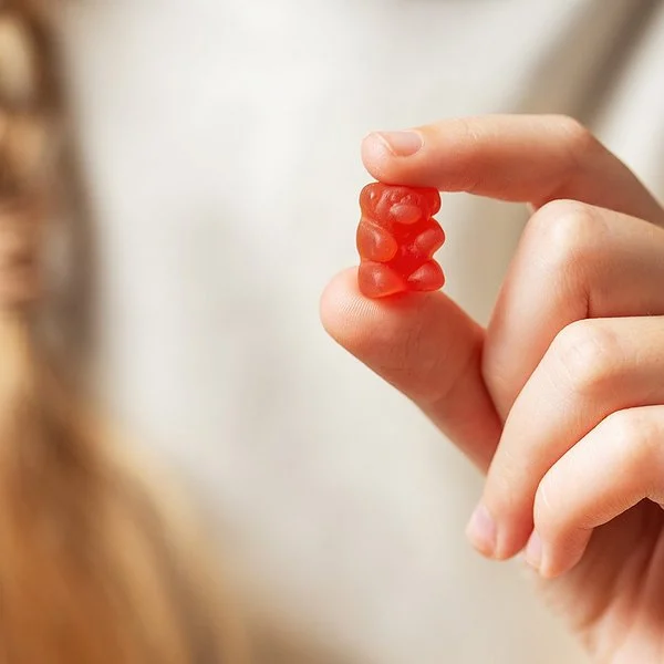 Im Vordergrund eine Kinderhand, die ein rotes Gummibärchen zwischen Zeigefinger und Daumen hält. Im Hintergrund in der Unsch