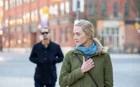 Eine blonde Frau läuft über einen öffentlichen Platz und blickt sorgenvoll über ihre Schulter. Hinter der Frau geht ein schwarz gekleideter Mann mit einer Sonnenbrille.