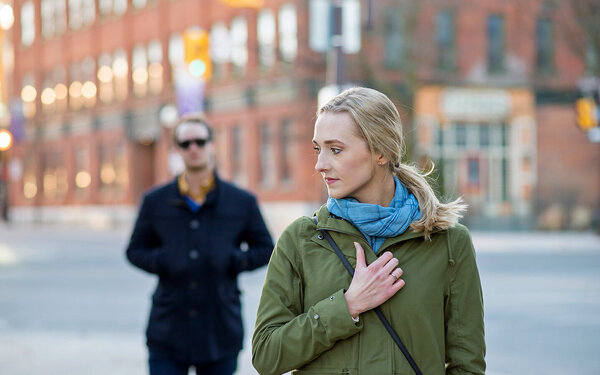 Eine blondhaarige Frau blickt über ihre Schulter, während sie die linke Hand auf ihrer Brust hält. Hinter der Frau geht ein schwarz gekleideter Mann.