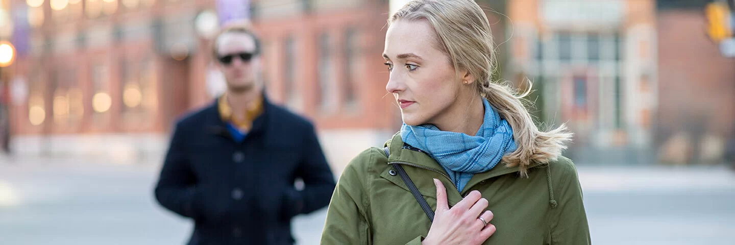 Eine blonde Frau läuft über einen öffentlichen Platz und blickt sorgenvoll über ihre Schulter. Hinter der Frau geht ein schwarz gekleideter Mann mit einer Sonnenbrille.